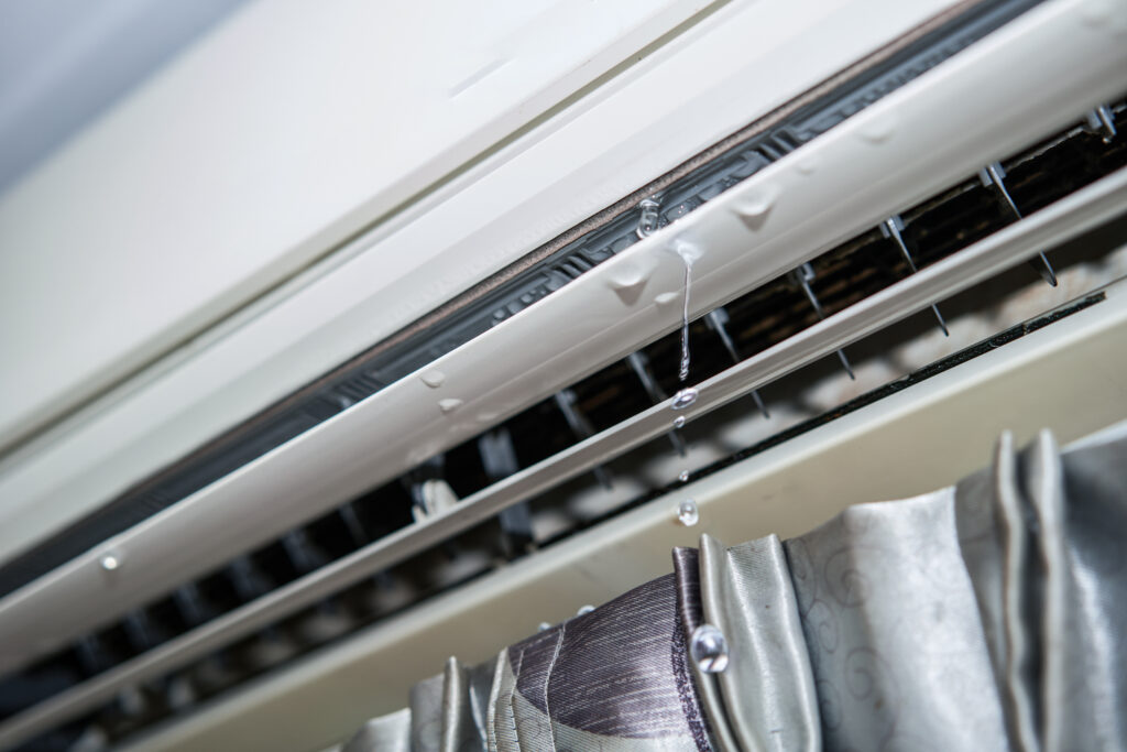 A close-up of a ductless air conditioning unit above a curtain with droplets of refrigerant leaking from the vents.