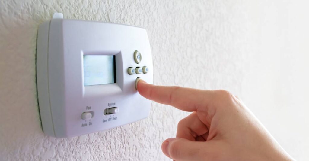 A person uses their finger to press the down button on a programmable thermostat installed on a textured white wall.