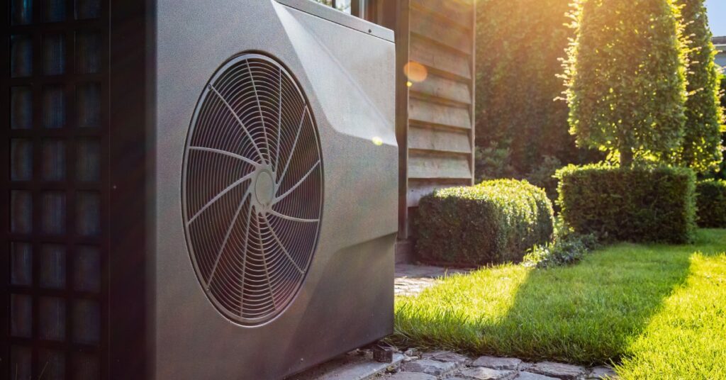 A heat pump with a dark gray exterior stands outside a home with the rays of the sun shining on it and the shrubs around it.