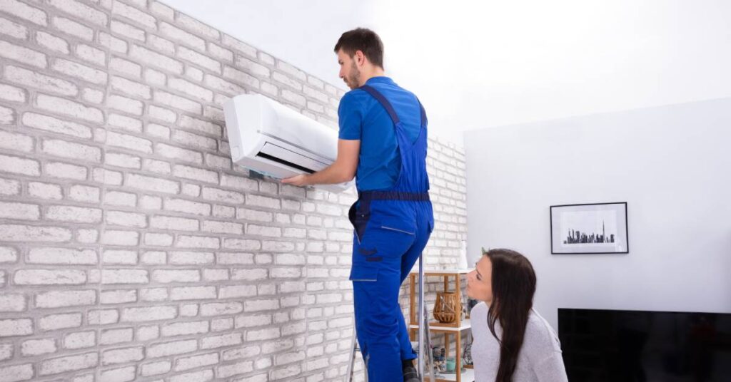 A man in a blue shirt and overalls stands on a ladder as he lifts an air conditioner onto the wall while a woman watches.