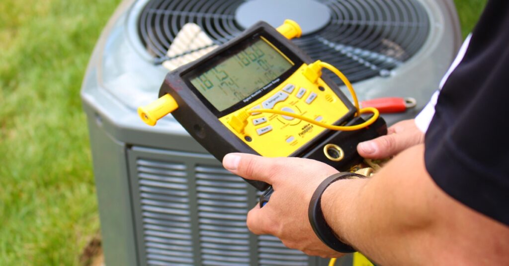 A person using a yellow and black device to test the electrical measurements of an air conditioner outside on a grassy lawn.