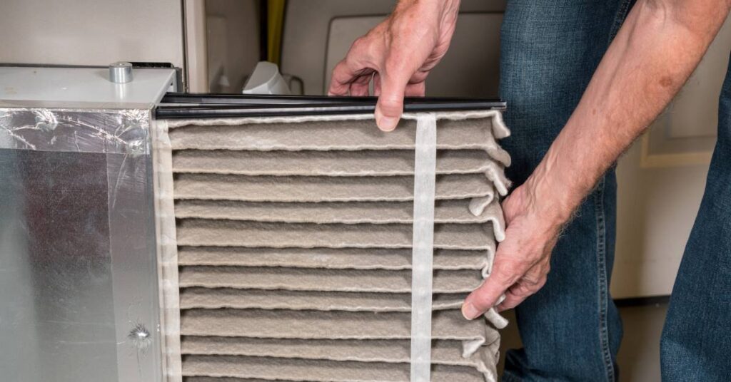 A person wearing jeans removes an air filter from a furnace. Layers of dirt and dust line the filter.