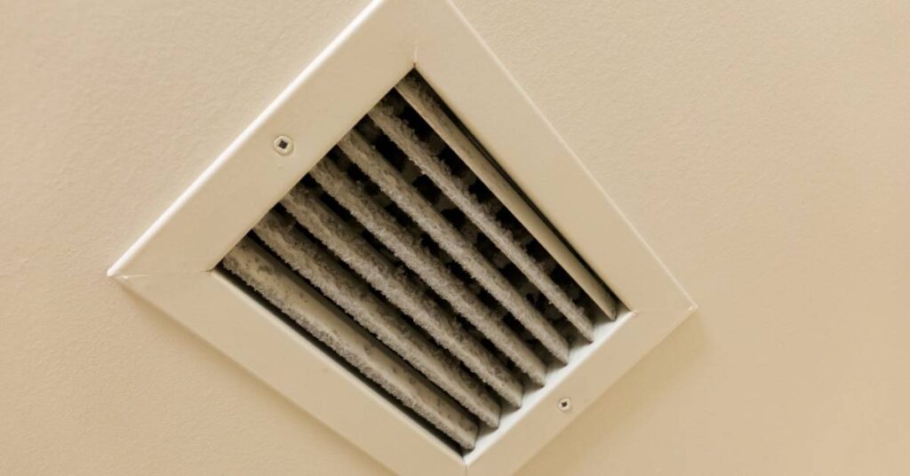 The opening of a ventilation duct in a white ceiling. Large clumps of dust line the rungs of the vent's opening.