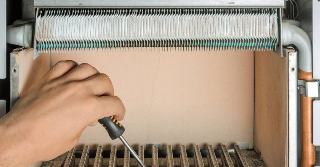 A person uses a screwdriver to adjust a component in between the rungs of a dirty gas burner showing signs of damage.