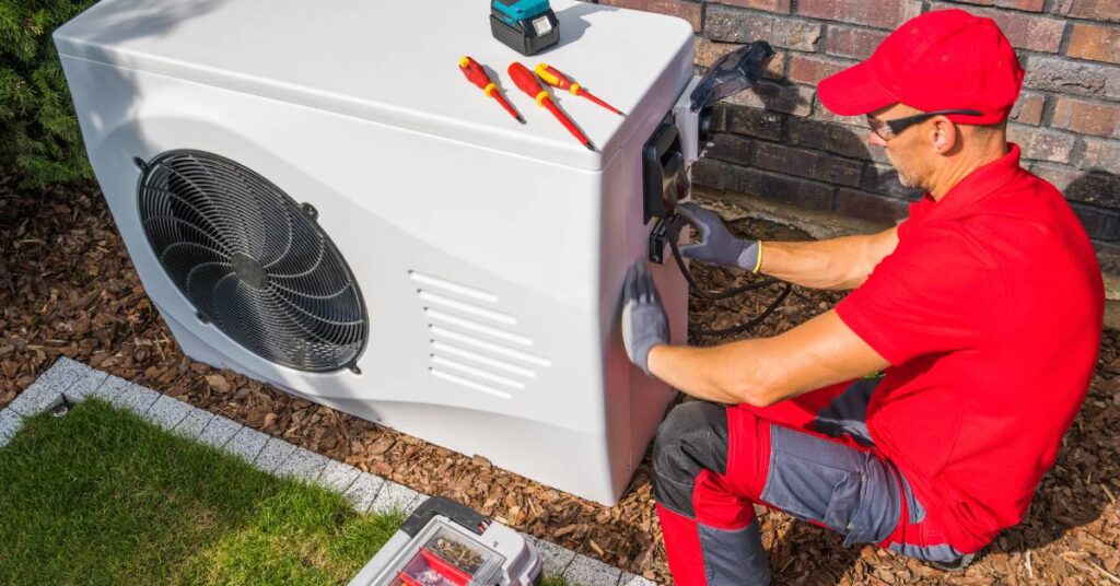 A man in a red shirt and black-and-red pants uses a variety of tools to work on the heat pump outside a brick building.