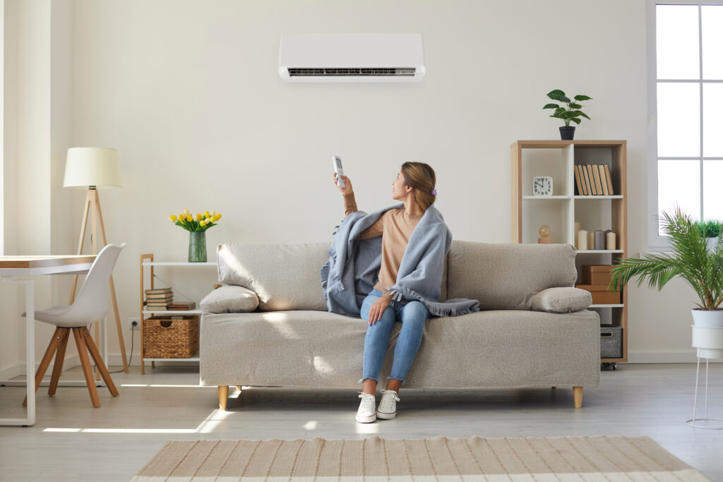 A woman with a blanket draped over her shoulders turns around on a couch to use the remote to control the air conditioner.