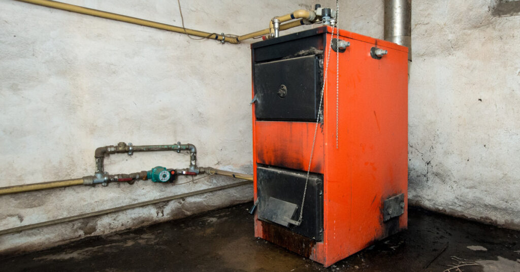 An old, orange furnace in the corner of a room with a patch of dark residue around it on the concrete floor.