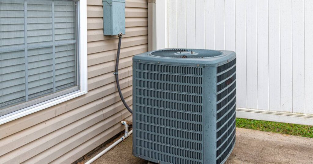 A central air-conditioning unit connected to a panel on the side of a house with beige shingles next to a white fence.