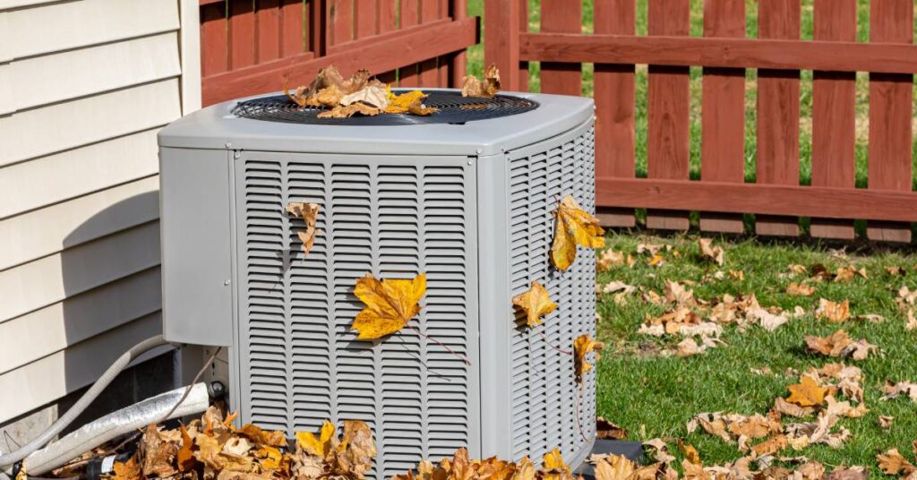 An air conditioner unit in the fenced-in yard of a house with beige siding. Brown leaves are lodged in the spaces of the unit.