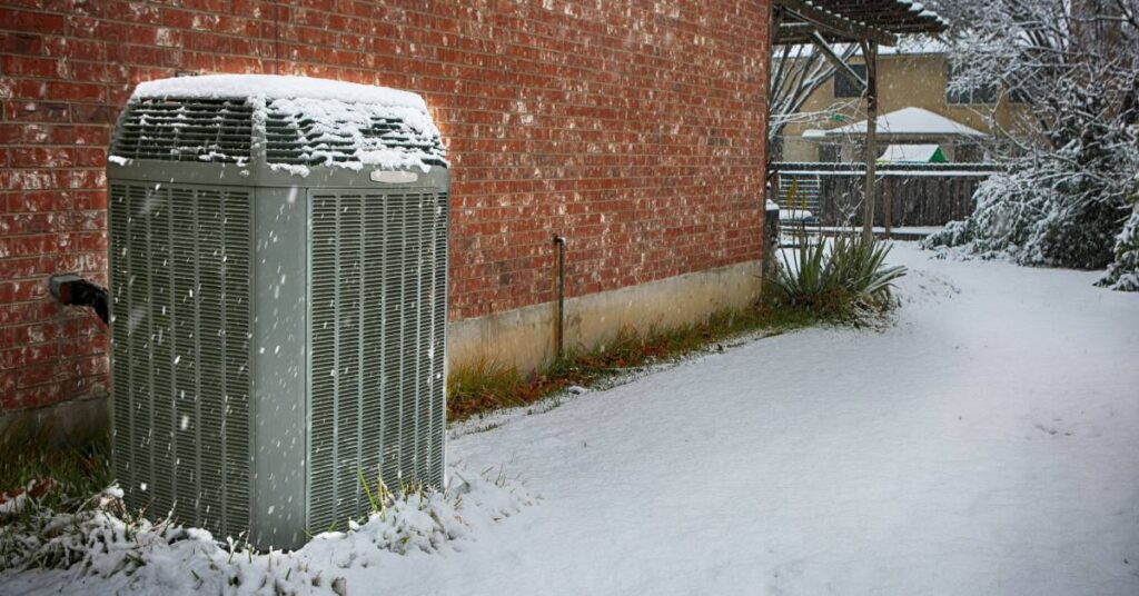 An air conditioner unit outside a brick house sits on the lawn. Snow is covering its top and surrounding it.